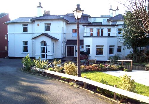 A large, white house with multiple storeys and a welcoming garden at the front serves as an inviting Wirral care home. There’s a lamp post and path leading gracefully to the entrance, embodying the warmth and community spirit of Abbeyfield.