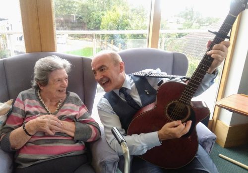 An elderly woman smiles at a man in a suit playing a guitar as they sit together on armchairs in the cosy Abbeyfield care home. Behind them, a window offers a serene outdoor view, enhancing their tranquil respite moment.