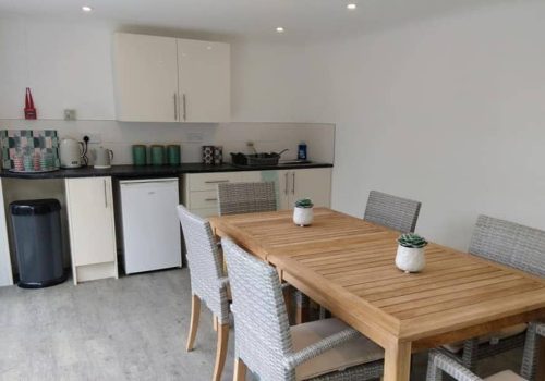 A cosy kitchen in the Wirral care home features white cupboards, a small fridge, and worktop appliances. A wooden dining table with six wicker chairs invites residents to gather, adorned with small potted plants for a touch of greenery.