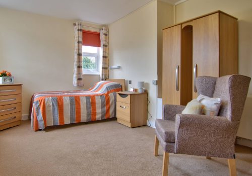 A tidy bedroom in a Wirral care home features a single bed with orange striped bedding, a wooden wardrobe, and a small side table with a teapot. A chest of drawers and an armchair near the window with curtains complete the comforting setup for assisted living.