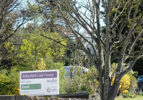 A sign for Abbeyfield Lear House with a CQC rating Good stands proudly on a stone wall, surrounded by trees and greenery, marking this welcoming residential care home.