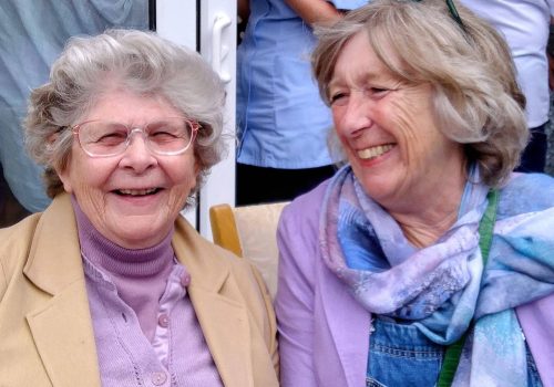 Two older women with grey hair, wearing spectacles and light-coloured clothes, are smiling warmly as they sit together indoors at a cosy Abbeyfield residential care home.
