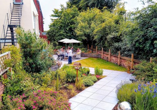 A landscaped garden with a meandering path, flowers, and shrubs. Four people are seated at a table beneath a white parasol in the background of this tranquil care home. A metal sphere is in the foreground, reflecting the tranquillity often associated with Abbeyfield and Wirral Care Home settings.