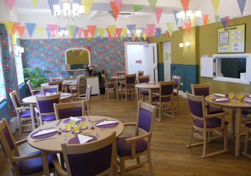 A colourful dining room in the Wirral care home features round tables laid with purple serviettes and glasses, while bright bunting hangs from the ceiling. The walls are adorned with floral and mustard-yellow accents, creating a welcoming space for residents enjoying respite care.