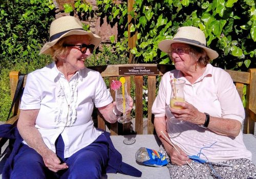 Two elderly women wearing hats sit outdoors on a bench at Abbeyfield Hoylake & West Kirby, smiling and holding drinks with straws and umbrellas. One has a knitting project beside her. A plaque on the bench is visible, adding a touch of charm to this tranquil scene.