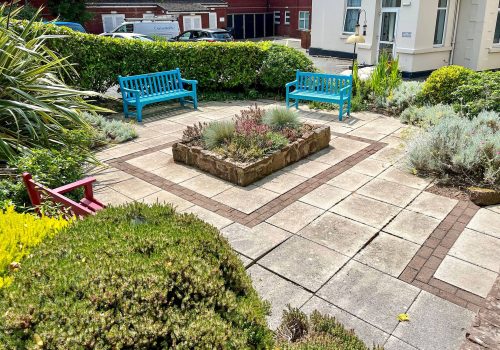 A small paved courtyard at the residential care home features two blue benches and one red bench. A central square planter is flanked by a variety of shrubs and greenery along the edges, providing a peaceful retreat for residents of the abbeyfield community.
