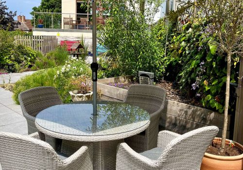 On the patio of our assisted care home, youll find a round glass table with five wicker chairs shaded by a large beige umbrella. Surrounded by greenery and landscaping, this tranquil spot under the clear blue sky provides a peaceful retreat.