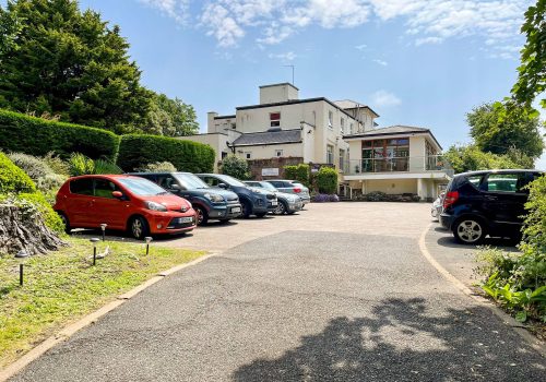 A car park with several cars, adjacent to a white, multi-storey residential care home surrounded by greenery on a sunny day.
