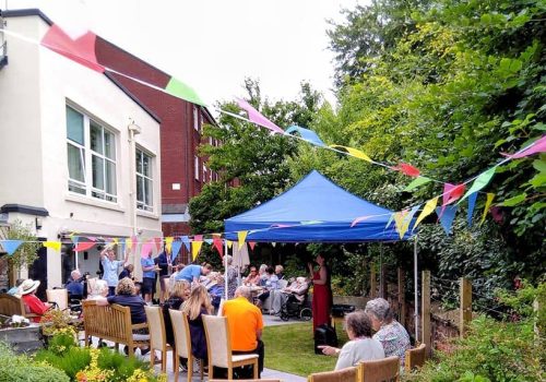 A group of people gather in a garden under a blue canopy, with colourful bunting overhead at the Wirral Care Home. Some are seated on chairs, while others stand or chat near a brick building, enjoying the lively atmosphere and camaraderie that comes with assisted care living.